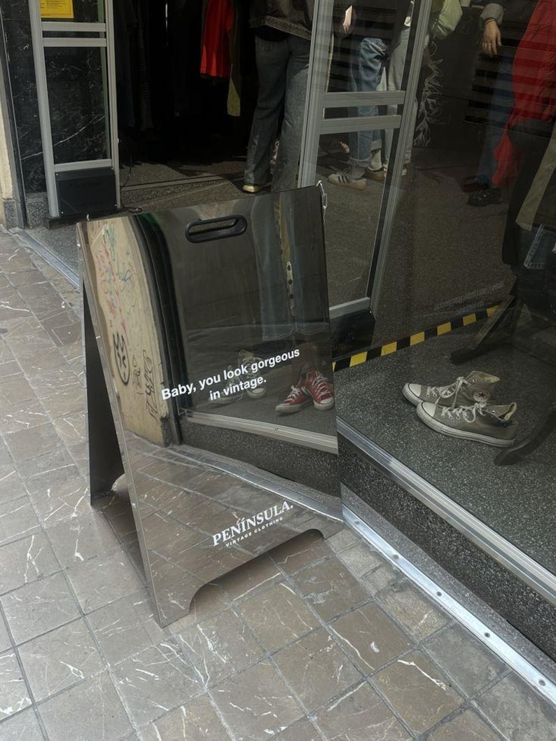 May include: A silver, reflective A-frame sidewalk sign with a handle cutout at the top. The sign features the white text "Baby, you look gorgeous in vintage." and "PEN&Iacute;NSULA VINTAGE CLOTHING" printed on the surface, reflecting the surrounding storefront.