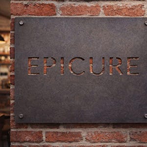 K&ouml;nnte beinhalten: Ein rechteckiges, dunkelgraues Schild mit dem ausgeschnittenen Wort "EPICURE", das an einer roten Backsteinmauer befestigt ist. Das Schild ist mit vier silbernen Schrauben befestigt. Der Hintergrund zeigt ein verschwommenes Interieur mit Tischen und H&auml;ngelampen.