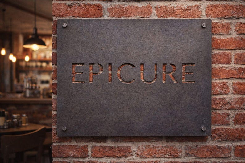K&ouml;nnte beinhalten: Ein rechteckiges, dunkelgraues Schild mit dem ausgeschnittenen Wort "EPICURE", das an einer roten Backsteinmauer befestigt ist. Das Schild ist mit vier silbernen Schrauben befestigt. Der Hintergrund zeigt ein verschwommenes Interieur mit Tischen und H&auml;ngelampen.