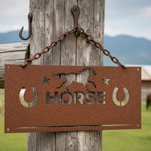K&ouml;nnte beinhalten: Ein rechteckiges, rostiges Metallschild mit dem ausgeschnittenen Wort "HORSE", einer Pferdesilhouette, Sternen und Hufeisen. Das Schild h&auml;ngt an einer Kette an einem Holzpfosten, mit grasenden Pferden im Hintergrund.