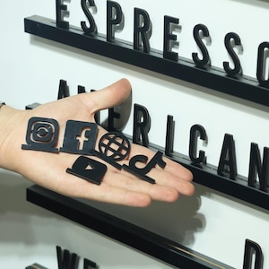 May include: A collection of small, black, flat social media icons including Instagram, Facebook, a globe, YouTube, and TikTok, held in a palm. In the background, a white letter board features black plastic letters spelling ESPRESSO and AMERICAN.