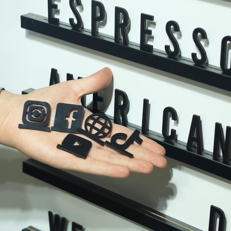 May include: A collection of small, black, flat social media icons including Instagram, Facebook, a globe, YouTube, and TikTok, held in a palm. In the background, a white letter board features black plastic letters spelling ESPRESSO and AMERICAN.