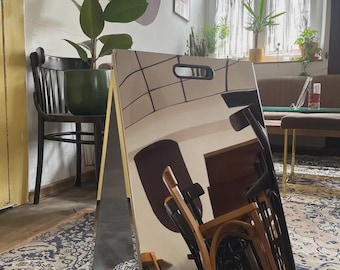 May include: A freestanding A-frame sign with a highly reflective, mirror-like silver surface. The sign features a rectangular cutout handle at the top center. The polished surface reflects the surrounding room, including chairs and indoor plants.