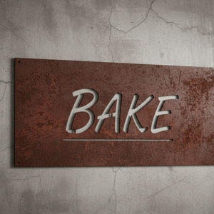 K&ouml;nnte beinhalten: Rechteckiges, rostbraunes Metallschild mit dem Wort "BAKE" in wei&szlig;en, stilisierten Buchstaben ausgeschnitten. Das Schild ist an einer strukturierten, grauen Betonwand angebracht, was auf ein K&uuml;chen- oder B&auml;ckereithema hindeutet.