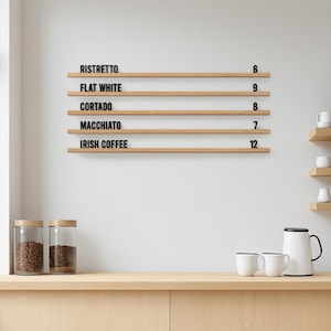 May include: A wall-mounted wooden menu board featuring five horizontal shelves with black lettering. The text reads RISTRETTO 6, FLAT WHITE 9, CORTADO 8, MACCHIATO 7, and IRISH COFFEE 12. The board is made of light-toned wood with a natural grain finish.