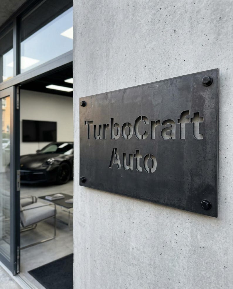 K&ouml;nnte beinhalten: Ein rechteckiges, dunkles Metallschild mit den ausgeschnittenen Worten "TurboCraft Auto". Das Schild ist an einer strukturierten, grauen Betonwand mit vier sichtbaren Schrauben befestigt. Durch eine offene T&uuml;r ist ein schwarzes Auto zu sehen.