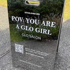 May include: A black reflective A-frame sidewalk sign with white text reading ESTABLISHED 2015, POV: YOU ARE A GLO GIRL, GLO SALON, LASHES | SUPPLY | ACADEMY, and a QR code with the text BOOK USING OUR QR CODE OR COME VISIT US INSIDE with an arrow.
