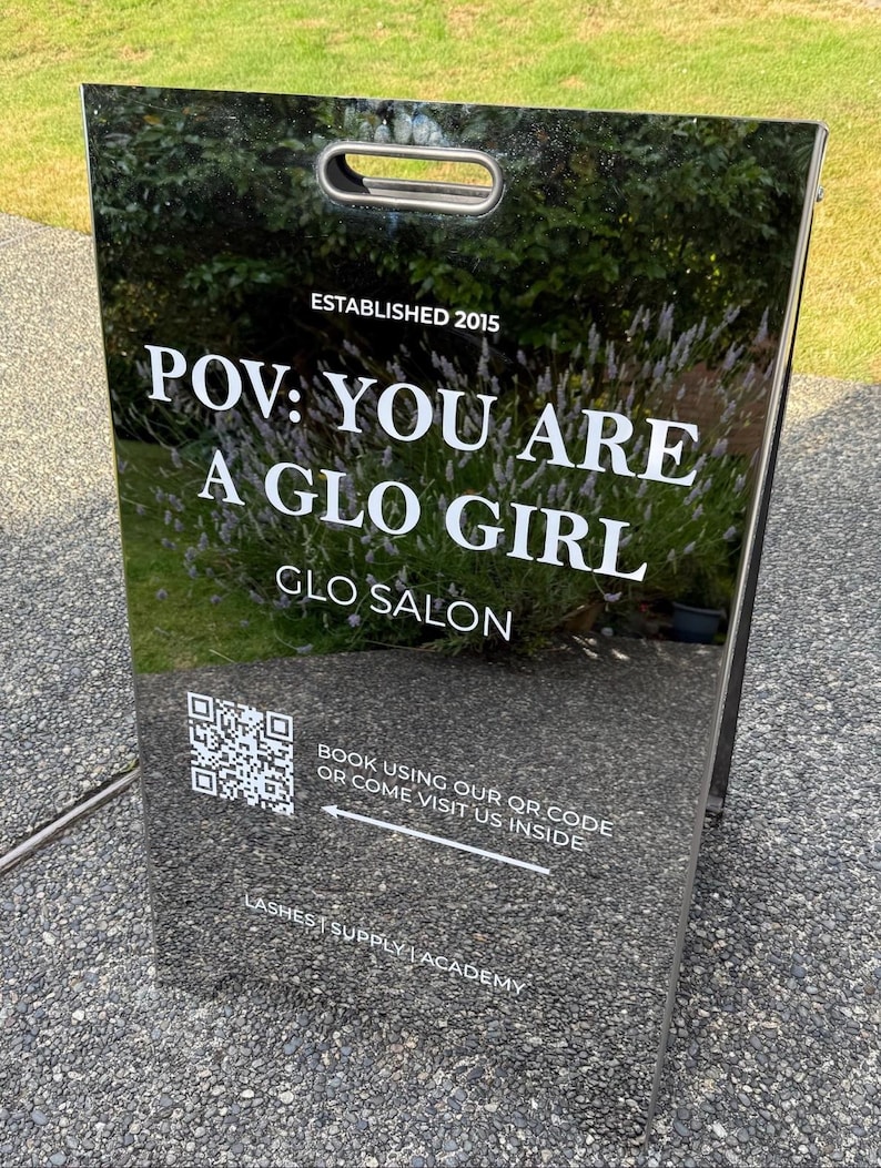 May include: A black reflective A-frame sidewalk sign with white text reading ESTABLISHED 2015, POV: YOU ARE A GLO GIRL, GLO SALON, LASHES | SUPPLY | ACADEMY, and a QR code with the text BOOK USING OUR QR CODE OR COME VISIT US INSIDE with an arrow.