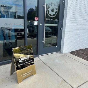 May include: A gold-colored A-frame sidewalk sign with a reflective surface featuring the white text Gilded signature handmade artisan soy candles, a website URL, and a social media handle, positioned on a concrete sidewalk outside a glass door.