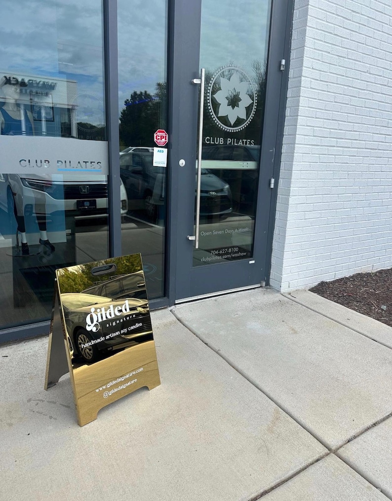 May include: A gold-colored A-frame sidewalk sign with a reflective surface featuring the white text Gilded signature handmade artisan soy candles, a website URL, and a social media handle, positioned on a concrete sidewalk outside a glass door.