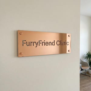K&ouml;nnte beinhalten: Ein rechteckiges, kupferfarbenes Schild mit den ausgeschnittenen Worten "FurryFriend Clinic". Das Schild ist mit vier sichtbaren Schrauben an einer hellen Wand befestigt. Das Schild befindet sich in einem hellen Raum.