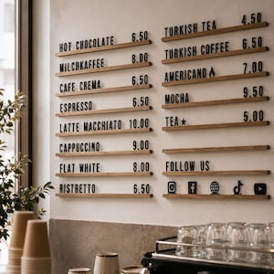 Puede incluir: Un tablero de menú montado en la pared con rieles de madera horizontales, letras negras y precios numéricos. El menú incluye artículos como Hot Chocolate, Milchkaffee, Cafe Crema, Espresso, Latte Macchiato, Cappuccino, Flat White, Ristretto, Turkish Tea, Turkish Coffee, Americano, Mocha y Tea.