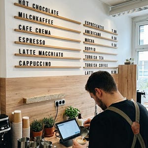May include: A cafe menu board with black lettering on horizontal wooden rails mounted to a white wall, listing items like Hot Chocolate, Milchkaffee, Cafe Crema, Espresso, Latte Macchiato, Cappuccino, Espreslatte, Americano, Mocha, Doppio, Turkish Tea, Turkish Coffee, and Latte.