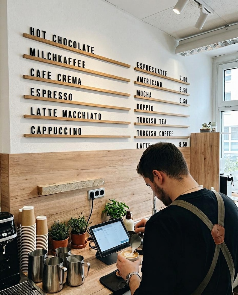 May include: A cafe menu board with black lettering on horizontal wooden rails mounted to a white wall, listing items like Hot Chocolate, Milchkaffee, Cafe Crema, Espresso, Latte Macchiato, Cappuccino, Espreslatte, Americano, Mocha, Doppio, Turkish Tea, Turkish Coffee, and Latte.
