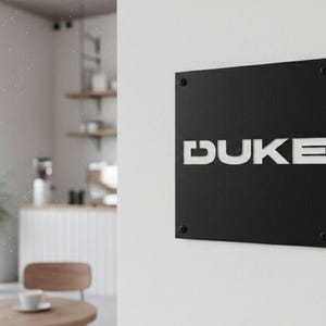 K&ouml;nnte beinhalten: Ein schwarzes quadratisches Schild mit dem wei&szlig;en Wort "DUKE" an einer wei&szlig;en Wand befestigt. Das Schild ist mit vier kleinen, dunklen Schrauben befestigt. Der Hintergrund zeigt eine Caf&eacute;-Umgebung mit Tisch, Stuhl und Pflanze.