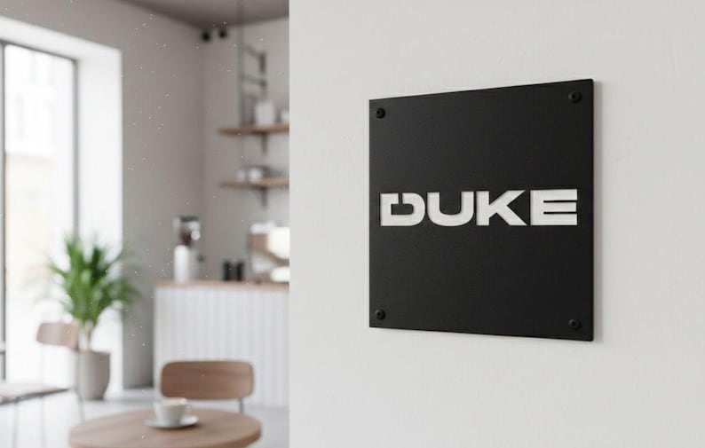 K&ouml;nnte beinhalten: Ein schwarzes quadratisches Schild mit dem wei&szlig;en Wort "DUKE" an einer wei&szlig;en Wand befestigt. Das Schild ist mit vier kleinen, dunklen Schrauben befestigt. Der Hintergrund zeigt eine Caf&eacute;-Umgebung mit Tisch, Stuhl und Pflanze.