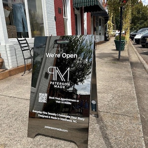 May include: A reflective A-frame sidewalk sign with the text We're Open, Peterson Made, and a QR code. It includes the phrases Book Your Appointment or Visit Us Inside, Everyday Fine Jewelry, Celebrate Your Story, Designed & Made in Matthews, NC, and petersonmade.com.