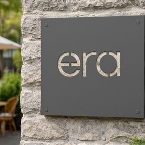K&ouml;nnte beinhalten: Ein dunkelgraues quadratisches Schild mit dem Wort "era" in der Mitte ausgeschnitten, an einer Steinmauer befestigt. Das Schild ist mit vier kleinen silbernen Schrauben befestigt. Der Hintergrund zeigt eine verschwommene Au&szlig;enszene mit Terrasse und B&auml;umen.