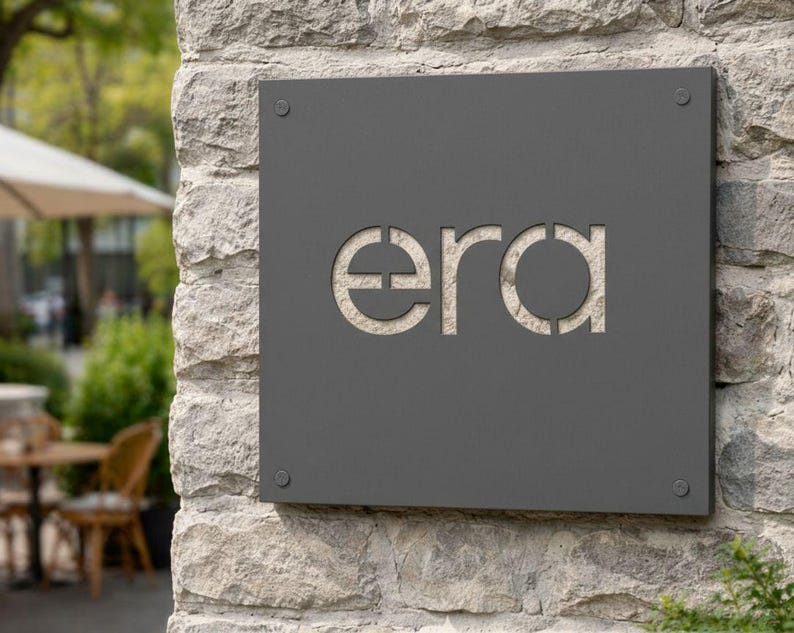 K&ouml;nnte beinhalten: Ein dunkelgraues quadratisches Schild mit dem Wort "era" in der Mitte ausgeschnitten, an einer Steinmauer befestigt. Das Schild ist mit vier kleinen silbernen Schrauben befestigt. Der Hintergrund zeigt eine verschwommene Au&szlig;enszene mit Terrasse und B&auml;umen.