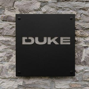 K&ouml;nnte beinhalten: Ein schwarzes quadratisches Schild mit dem ausgeschnittenen Wort "DUKE", montiert an einer Steinmauer. Das Schild ist mit vier dunklen Schrauben befestigt, und die Buchstaben sind in einer serifenlosen Schrift.