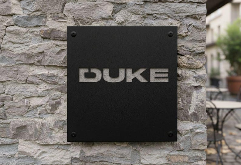 K&ouml;nnte beinhalten: Ein schwarzes quadratisches Schild mit dem ausgeschnittenen Wort "DUKE", montiert an einer Steinmauer. Das Schild ist mit vier dunklen Schrauben befestigt, und die Buchstaben sind in einer serifenlosen Schrift.