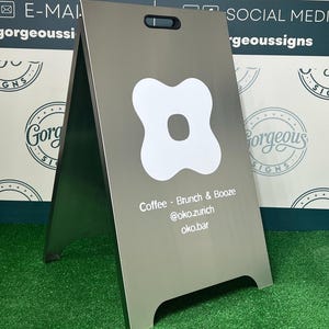 May include: A silver metal A-frame sidewalk sign with a handle cutout at the top. The sign features a white four-lobed graphic and white text that reads: Coffee - Brunch & Booze, @oko.zurich, and oko.bar, centered on the front panel.