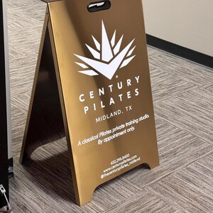 May include: A gold-colored A-frame sign with the text "CENTURY PILATES MIDLAND, TX" in white. The sign also includes contact information and the phrase "A classical Pilates private training studio. By appointment only."