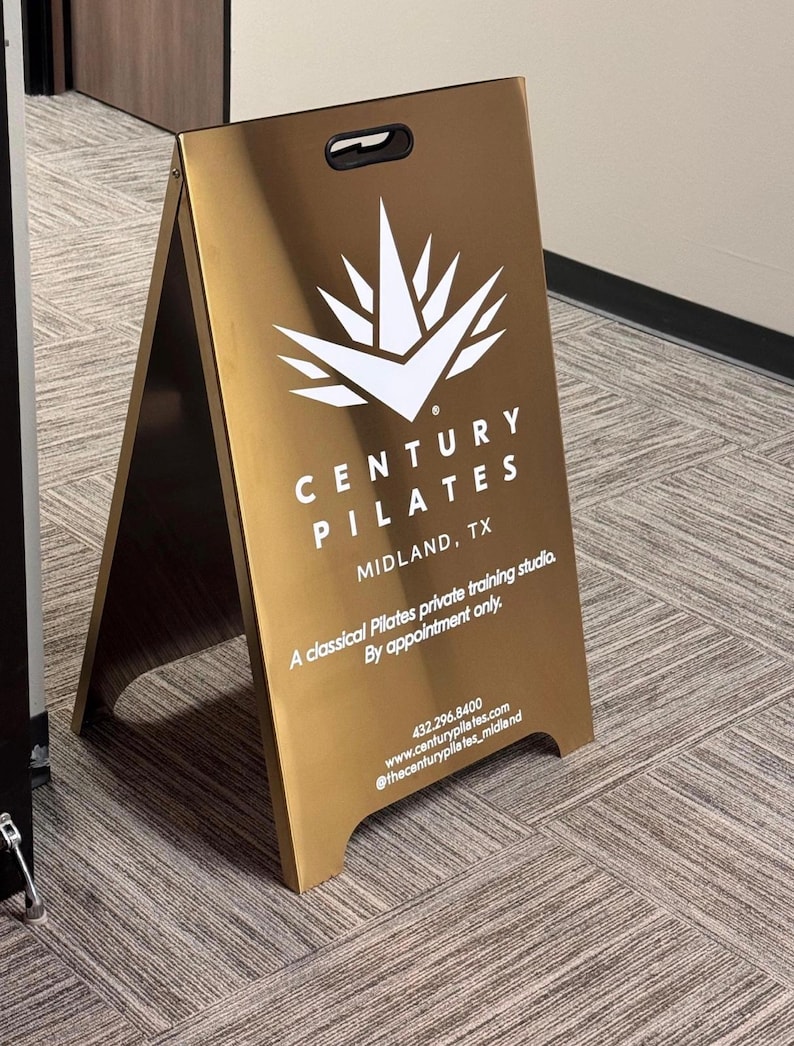 May include: A gold-colored A-frame sign with the text "CENTURY PILATES MIDLAND, TX" in white. The sign also includes contact information and the phrase "A classical Pilates private training studio. By appointment only."
