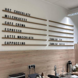 May include: A wall-mounted menu board featuring horizontal wooden rails with black letter tiles. The text reads: HOT CHOCOLATE, MILCHKAFFEE, CAFE CREMA, ESPRESSO, LATTE MACCHIATO, CAPPUCCINO, TURKISH TEA, and TURKISH COFFEE, arranged in two columns.