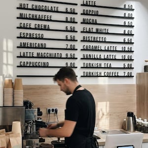 May include: A wall-mounted menu board with black horizontal rails displaying white text for coffee and tea items including Hot Chocolate 6.50, Latte 8.50, Doppio 8.00, Mocha 9.50, Flat White 8.00, Ristretto 6.50, Americano 7.00, and Turkish Coffee 7.00.