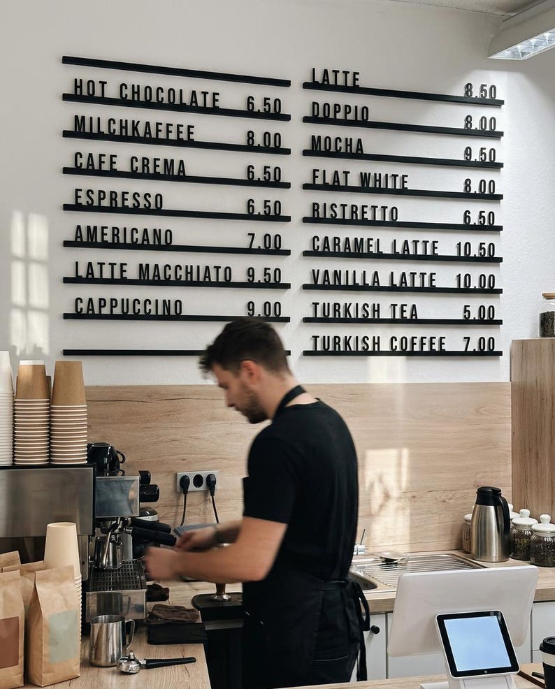 May include: A wall-mounted menu board with black horizontal rails displaying white text for coffee and tea items including Hot Chocolate 6.50, Latte 8.50, Doppio 8.00, Mocha 9.50, Flat White 8.00, Ristretto 6.50, Americano 7.00, and Turkish Coffee 7.00.