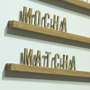 May include: Two horizontal light brown wooden shelves mounted on a wall, each featuring individual wooden letters standing upright. The top shelf displays the word MOCHA and the bottom shelf displays the word MATCHA in a sans-serif font.
