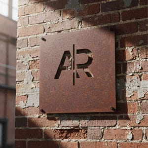 K&ouml;nnte beinhalten: Ein quadratisches, rostiges Metallschild mit den ausgeschnittenen Buchstaben "AR", das an einer verwitterten Backsteinmauer befestigt ist. Das Schild ist mit vier kleinen Schrauben befestigt. Die Backsteinmauer ist eine Mischung aus roten und braunen Ziegeln mit fehlendem M&ouml;rtel.