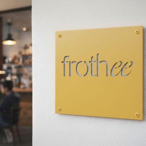 K&ouml;nnte beinhalten: Ein quadratisches, senfgelbes Schild mit dem Wort "frothee" aus Metall ausgeschnitten. Das Schild ist mit vier kleinen goldfarbenen Schrauben an einer wei&szlig;en Wand befestigt. Das Schild ist wahrscheinlich f&uuml;r ein Caf&eacute;.