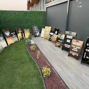May include: A collection of A-frame signs with various designs and text, including "CHID", "Best Pub of London", and "Barbershop Charisma". The signs are in different colors, including black, gold, and silver, and are displayed on a grassy area.
