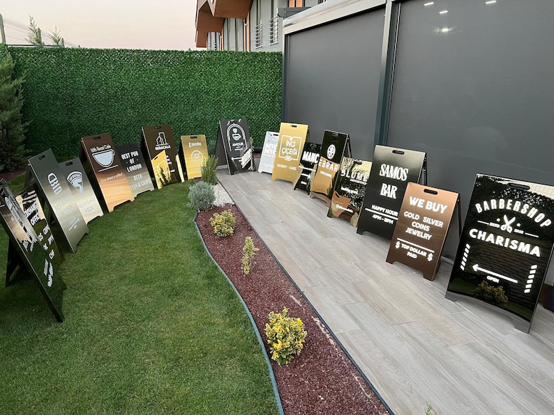 May include: A collection of A-frame signs with various designs and text, including "CHID", "Best Pub of London", and "Barbershop Charisma". The signs are in different colors, including black, gold, and silver, and are displayed on a grassy area.
