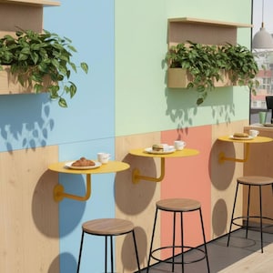 May include: Three wall-mounted yellow circular tables with matching stools featuring wooden seats and black metal frames. Each table holds a white cup and a small plate with food. Above each table is a wooden shelf containing green plants.