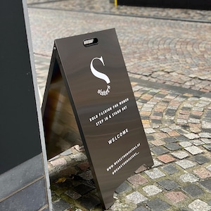 May include: A-frame sign with the word "sister's" in white script and a stylized "S" logo. The sign also includes the text "BOLD FASHION FOR WOMEN STEP IN & STAND OUT" and "WELCOME". The sign is brown and stands on a cobblestone street.
