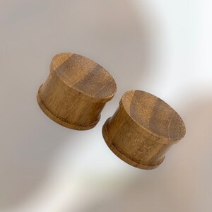 May include: Two wooden ear plugs with a light brown color and a textured surface. The plugs have a flared design and are likely used for body modification. The plugs are made of wood.