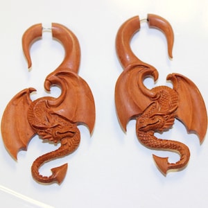 May include: A pair of wooden dragon ear gauges. The gauges are carved in the shape of a dragon with wings outstretched. The gauges are a reddish-brown colour.