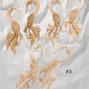 May include: Five cream-coloured, carved earrings with a hook design. Each earring features a curved hook, a wrapped section, and a detailed leaf-like design. The earrings are displayed on a white, textured surface.