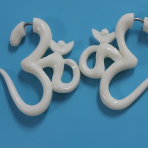 Può includere: Due orecchini in plastica bianca a forma di simbolo Om. Gli orecchini hanno la forma del simbolo Om e hanno un piccolo anello nella parte superiore per essere fissati a un piercing.