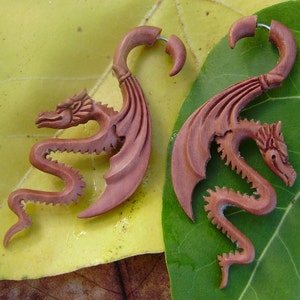 May include: A pair of wooden dragon ear gauges. The gauges are carved in a detailed dragon design with wings and a curled tail. The gauges are a reddish-brown colour.