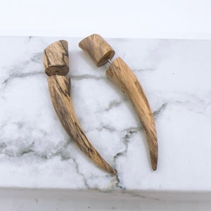 Op de afbeelding: Een paar houten oorpluggen met een uniek houtnerfpatroon. De pluggen zijn hoornvormig en hebben een zilveren metalen pen.