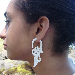 Op de afbeelding: Een witte, van bot gesneden oorbel met een bloemmotief. De oorbel is een groot, ingewikkeld ontwerp met een hoepel en een hangend bloemmotief.