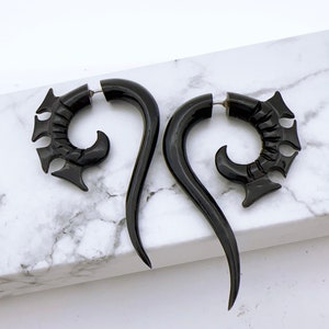 Op de afbeelding: Een paar zwarte hoorn oorpluggen met een gebogen, ingewikkeld ontwerp. De pluggen zijn gemaakt van natuurlijk hoorn en hebben een gepolijste afwerking.