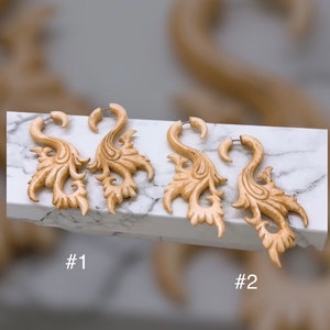 Op de afbeelding: Twee paar lichtgekleurde, gesneden houten oorbellen met een sierlijk, bloemmotief. Elke oorbel heeft een gebogen haak en een gedetailleerd, wervelend patroon. De oorbellen zijn gelabeld met #1 en #2.