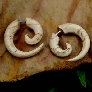 May include: A pair of wooden spiral earrings with a natural wood grain pattern. The earrings are made of light brown wood and have a silver metal post.