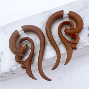 Op de afbeelding: Een paar bruine houten oorbellen met een gesneden, wervelend ontwerp. De oorbellen hebben een gebogen vorm met ingewikkelde details en een zilverkleurige pin voor piercing. De oorbellen worden gepresenteerd op een wit en grijs marmeren oppervlak.