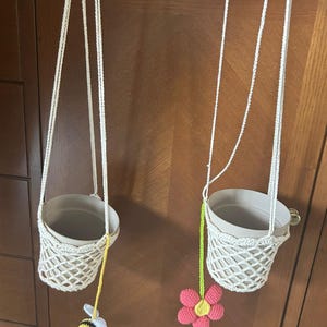 Peut inclure: Deux porte-plantes suspendus en macramé avec des pots beiges. Les supports sont en macramé blanc et sont suspendus par des cordons blancs. L'un a une abeille jaune et noire, l'autre une fleur rose et jaune.